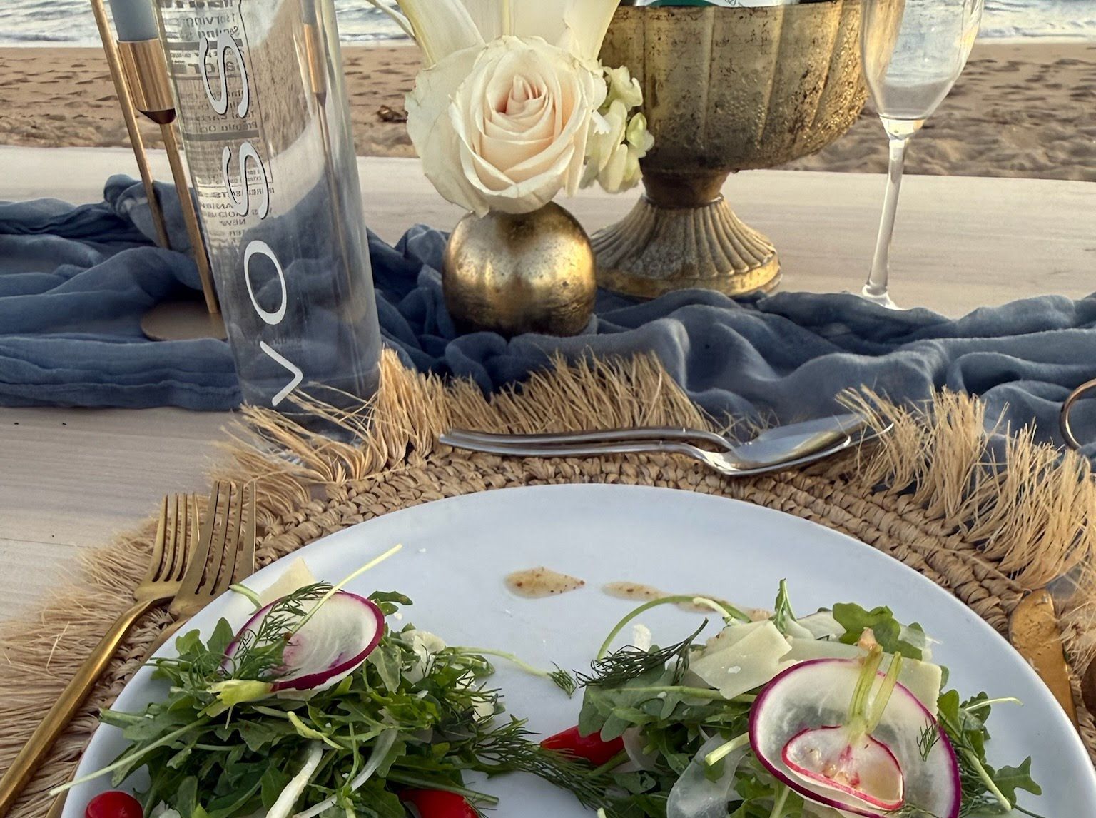A table detail on the beach: a white rose in a small gold vase, soft blue linen, a woven charger and a plated salad.