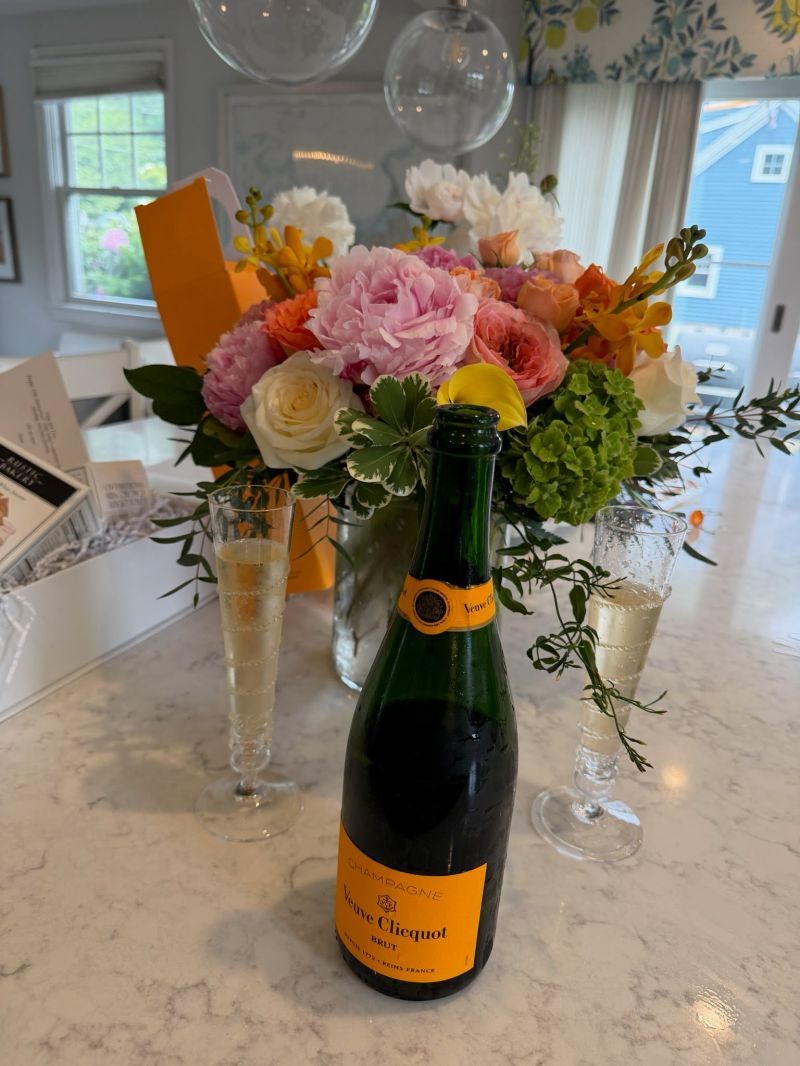 A bouquet of peonies, roses and garden flowers beside a bottle of champagne and two filled flutes on a white marble counter.
