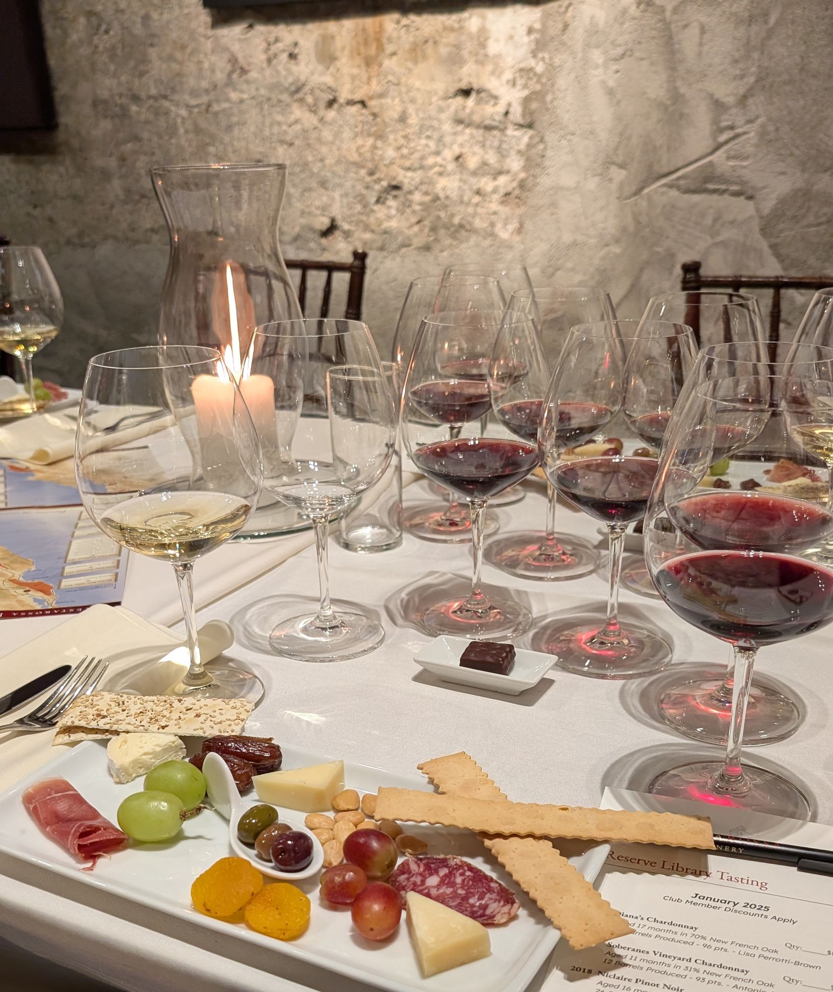 A long table set for a wine tasting in a stone-walled room, with rows of glasses, a lit candle and a board of cheese, fruit and charcuterie.